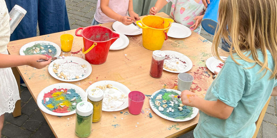 Kinder stehen um einen Tisch und gestalten auf Papptellern mit buntem Sand und Muscheln Landschaften