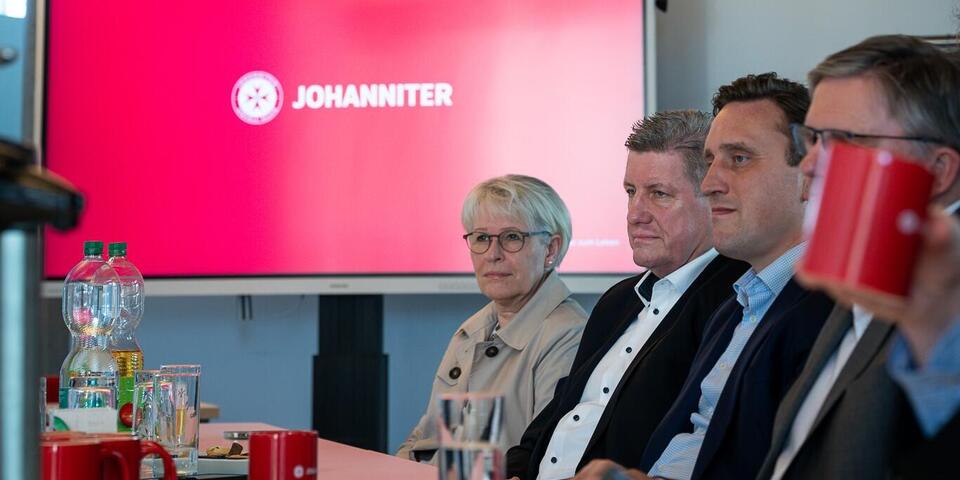 Menschen an einem Konferenztisch. Im Hintergrund ein großer Bildschirm auf dem der Schriftzug Johanniter mit roter Hintergrundfarbe zu sehen ist