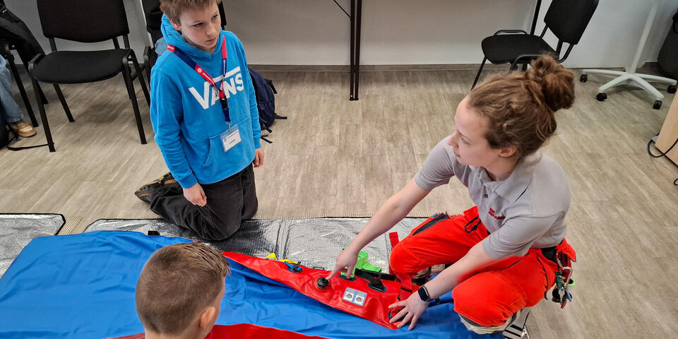Eine Frau in Einsatzkleidung kniet neben einer Vakuummatratze und spricht. Kinder schauen ihr zu.