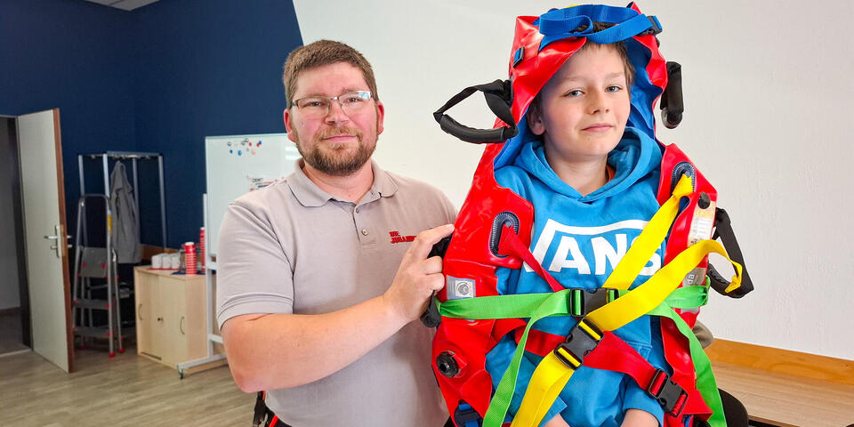 Ein Mann in Einsatzkleidung steht neben einem Jungen, der in einer Vakuummatratze eingewickelt ist.