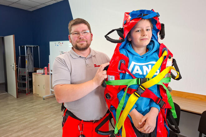 Ein Mann in Einsatzkleidung steht neben einem Jungen, der in einer Vakuummatratze eingewickelt ist.