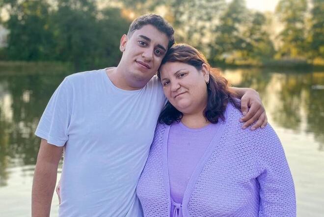 Ein junger Mann und seine Mama stehen Arm in Arm auf einem Steg vor einem ruhigen See. Beide lächeln leicht und blicken in die Kamera. Die Atmosphäre ist ruhig und herzlich, Bäume spiegeln sich im Wasser im Hintergrund.