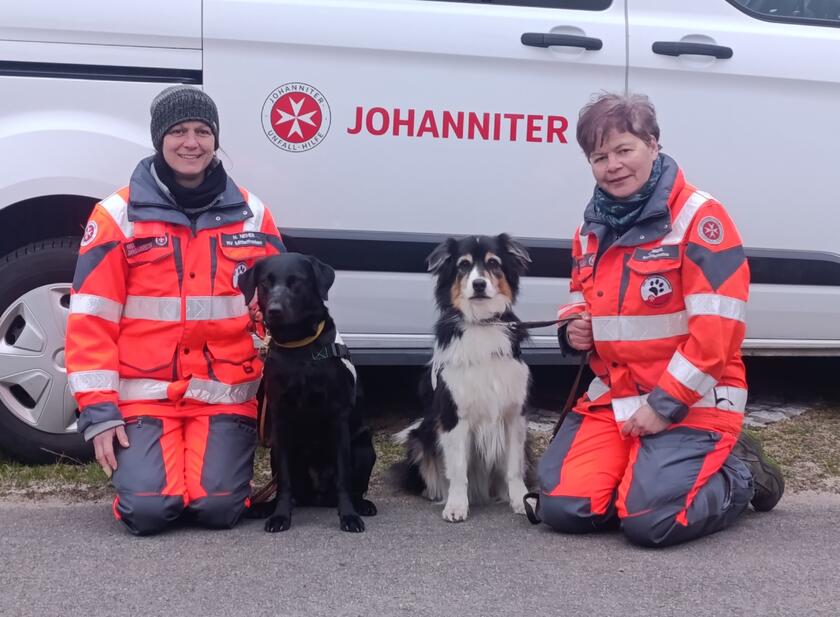 Die beiden Rettungshundeführerinnen in Einsatzkleidung knien mit ihren Hunden vor dem Fahrzeug der Johanniter.