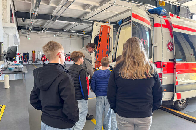 Ein Mann zieht eine Trage aus einem Rettungswagen. Kinder schauen ihm zu.