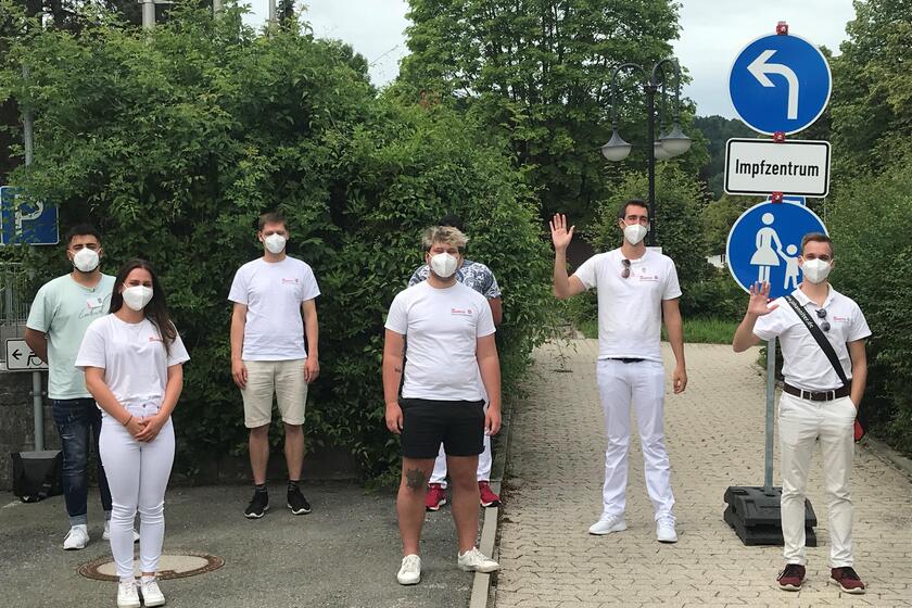 Mehrere junge Erwachsene mit Mund-Nasen-Schutz stehen vor einem Straßenschild mit der Aufschrift „Impfzentrum“, zwei von ihnen winken in die Kamera. Dabei halten sie alle den Mindestabstand ein.