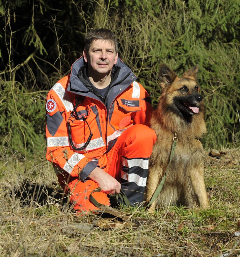 Ein Johanniter-Rettungshundeführer kniet in auffälliger Einsatzkleidung im Wald und hält seinen Deutschen Schäferhund an der Leine. Beide schauen aufmerksam und freundlich in die Kamera, bereit für ihren nächsten Einsatz.