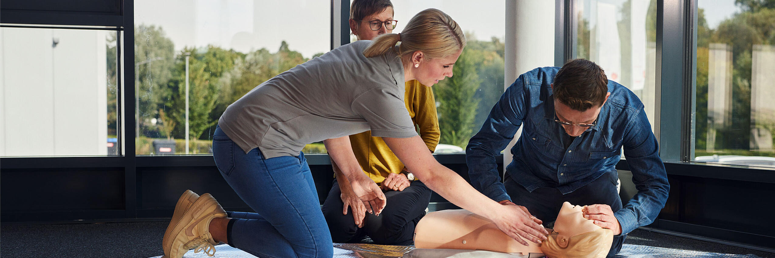 Eine Erste-Hilfe-Trainerin der Johanniter weist zwei Teilnehmende eines Erste-Hilfe-Kurses zur Reanimation an einer Übungspuppe an.