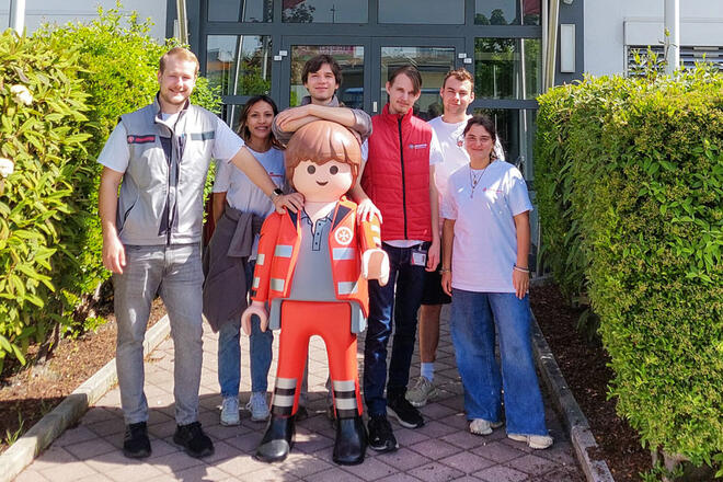 Mehrere junge Leute in Johanniter Kleidung stehen um ein großes Johanniter Playmobilmännchen in Einsatzkleidung.