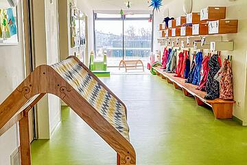 Heller Flur einer Kita mit bunten Garderobenhaken, niedrigen Sitzbänken und einem kleinen Spielhäuschen im Vordergrund.