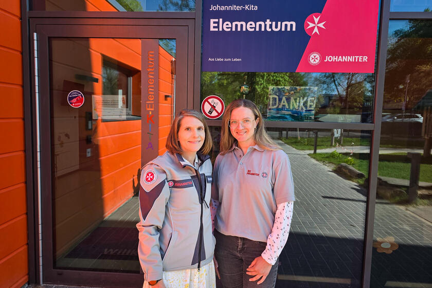 Zwei Frauen mit Johanniter-Oberbekleidung vor einer Glasfront mit einem Schild auf dem steht "Johanniter-Kita Elementum"