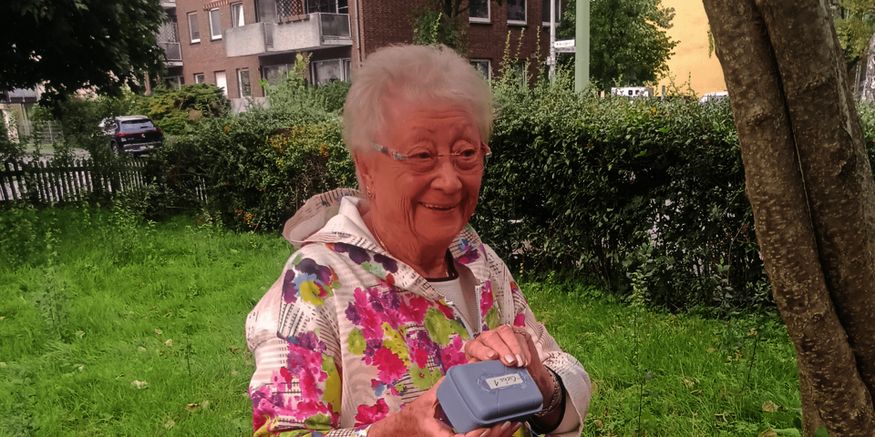 Eine Frau in einer geblümten Jacke steht auf einer Wiese und hält ein kleines graues Kästchen in der Hand.