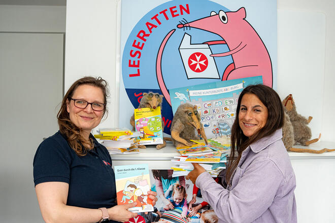 Zwei Frauen stehen vor mehreren Kinderbüchern, hinter ihnen ist das Logo der Johanniter Leseratten.
