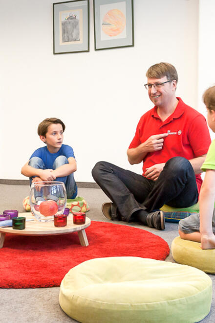Kinder und ein Betreuer sitzen auf Kissen um einen kleinen Tisch mit Kerzen und Dekoration.