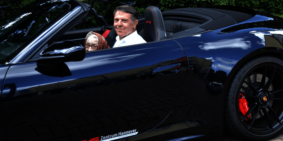 Eine weißhaarige Frau und ein größere Mann sitzen nebeneinander in einem schwarzen Auto mit offenem Verdeck.