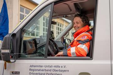 Johanniterin in orangefarbener Einsatzkleidung sitzt lächelnd am Steuer eines Einsatzfahrzeugs.