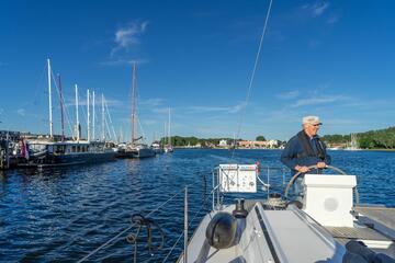 Ein Ehrenamtlicher steuert das Segelboot, im Hintergrund ist der Yachthafen von Travemünde zu sehen.