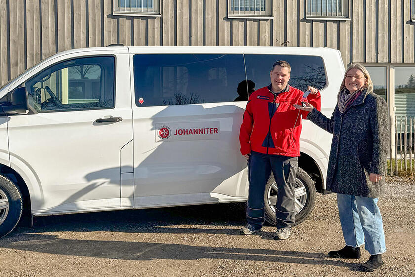 Zwei Erwachsene stehen vor einem weißen Johanniter-Transporter und halten gemeinsam einen Autoschlüssel vor einem Holzgebäude.
