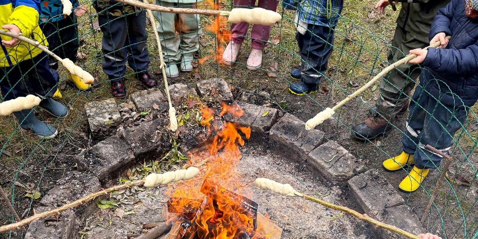 Ein Lagerfeuer. Über dem Feuer werden Äste mit Stockbrot gehalten.