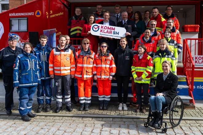 Eine große Gruppe von Menschen in Rettungsdienst- und Einsatzkleidung steht vor einem roten Feuerwehrauto und blickt in die Kamera.