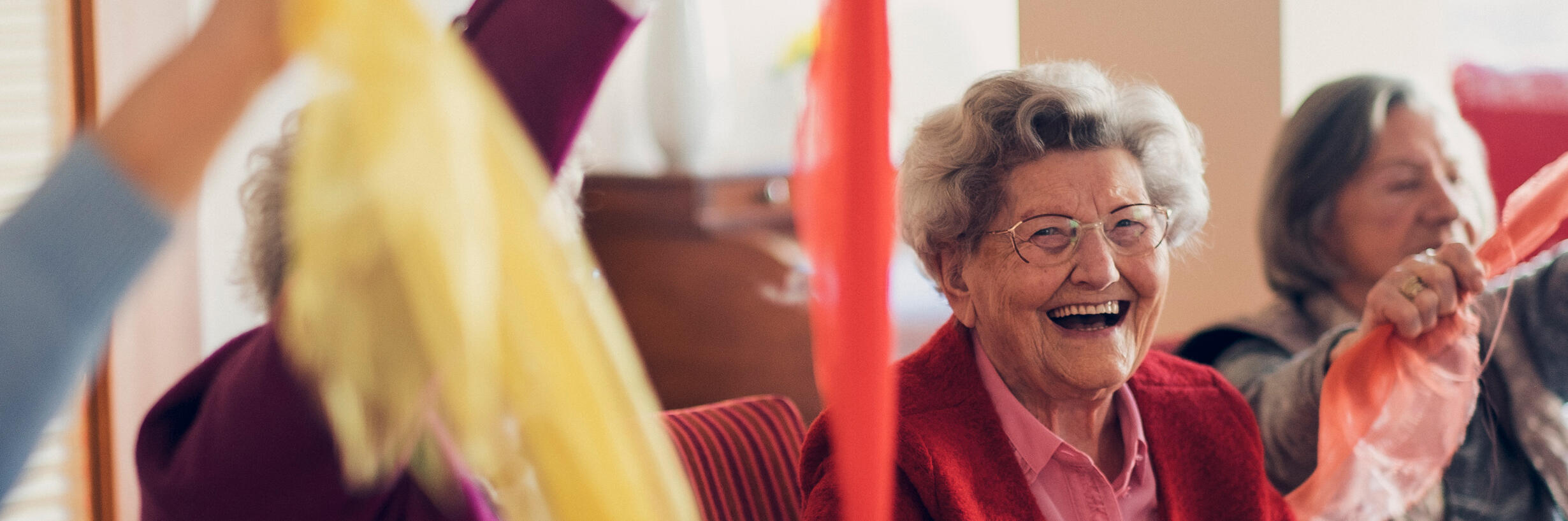Lachende auf dem Stuhl sitzende Seniorinnen bewegen sich spielend mit einem bunten Tuch