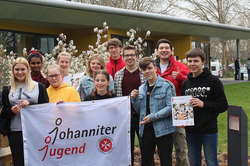 Ein Gruppenbild von Schüler*innen mit zwei jungen Erwachsenen. Im Vordergrund ein Johanniter-Jugend-Banner