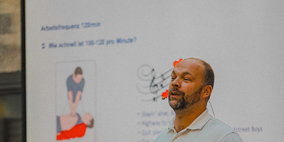 Ein Mann mit weißem Johanniter-Polo steht vor einem Bildschirm auf dem eine Grafik mit Herzdruckmassage zu sehen ist.