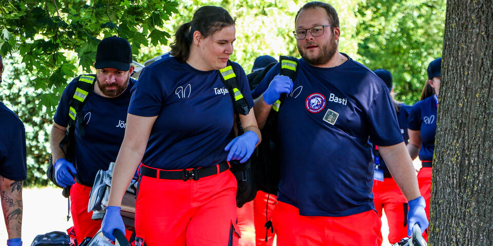 Mehrere Einsatzkräfte in Einsatzkleidung tragen unter Bäumen medizinische Geräte und Ausrüstung zu einem Einsatzort.
