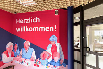 Ein großes Wandbild mit Illustrationen von Menschen verschiedenen Alters und der Aufschrift "Herzlich willkommen" begrüßt die Gäste im Begegnungsraum Liederbach.