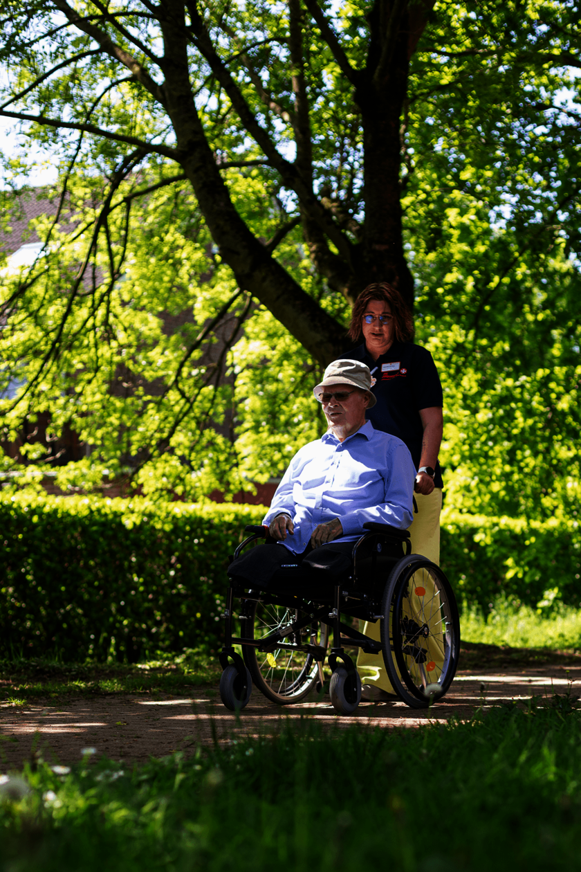 Pflegerin schiebt einen älteren Herrn im Rollstuhl auf einem schattigen Weg unter Bäumen.