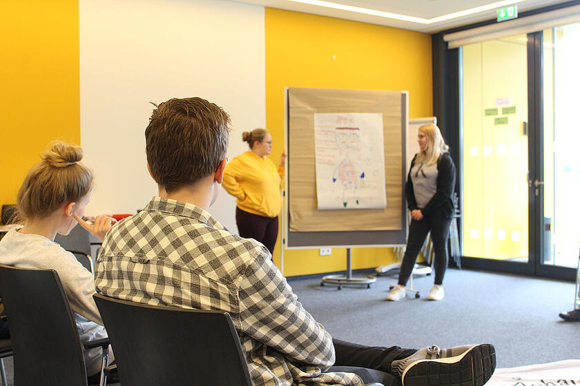 Zwei junge Menschen sitzen im Vordergrund auf Stühlen. Sie hören einem Mädchen zu, das im Hintergrund etwas an einer Pinnwand erklärt.