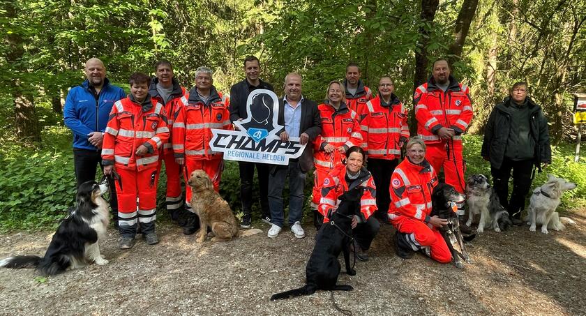 Gruppe von Einsatzkräften und weiteren Personen steht mit mehreren Hunden und einem Schild „Champs – Regionale Helden“ auf einem Waldweg.