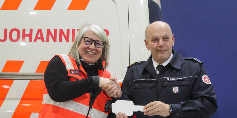 Ein Mann überreicht einer Frau in Johanniter-Einsatzbekleidung einen großen weißen Schlüssel aus Pappe.