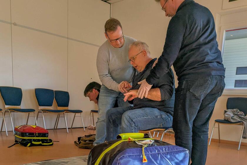 Zwei Männer kümmern sich um einen älteren Mann, der auf einem Stuhl sitzt und Probleme mit der Atmung hat. Ein weiterer Mann kniet über einem Rucksack.