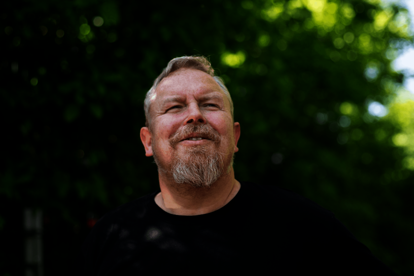 Das Porträt zeigt einen bärtigen Mann, der vor grüner Bepflanzung nach oben schaut