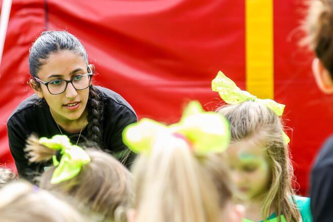 Junge Teamerin beugt sich zu einer Kindergruppe; Neon-Schleifen vor roter Plane.