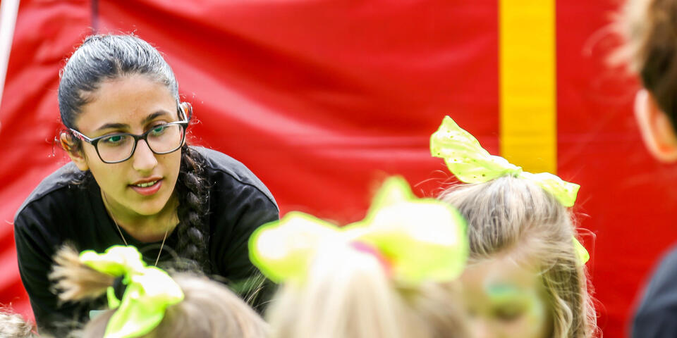 Junge Teamerin beugt sich zu einer Kindergruppe; Neon-Schleifen vor roter Plane.