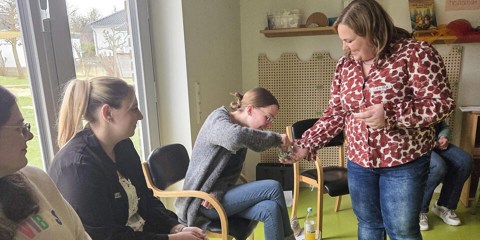 Drei Frauen sitzen auf Stühlen. Eine Frau geht mit einem Glas auf dem "Glück" steht, herum. Eine sitzenden Frau greift in das Glas.