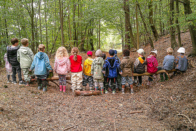 Kinder stehen an und sitzen auf einem querliegendem großen Ast im Wald