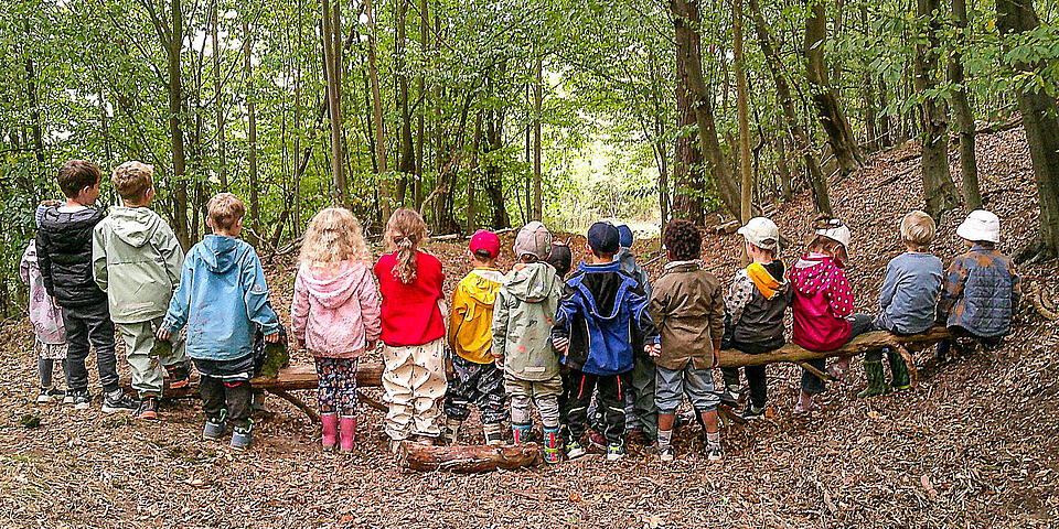 Kinder stehen an und sitzen auf einem querliegendem großen Ast im Wald