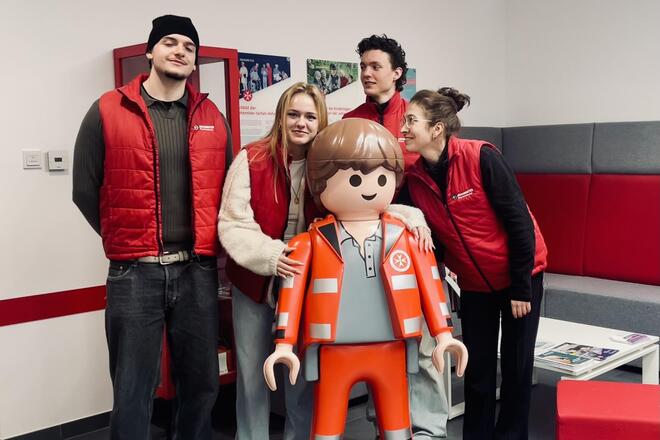 Auf dem Gruppenfoto zeigen sich die vier neuen Mitarbeitenden der Johanniter, die in den nächsten Wochen auf der Suche nach neuen Fördermitgliedern sind. Fröhlich lachend zeigen sie sich in einem hellen Raum mit einem großen Johanniter Playmobilmännchen.
