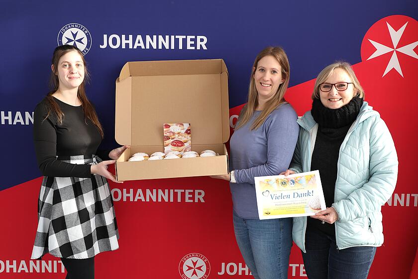 Vertreterin des Lions Clubs Regensburg Therese von Schwandorf mit zwei Mitarbeitenden der Johanniter in Ostbayern, die einen Karton Krapfen in die Kamera halten