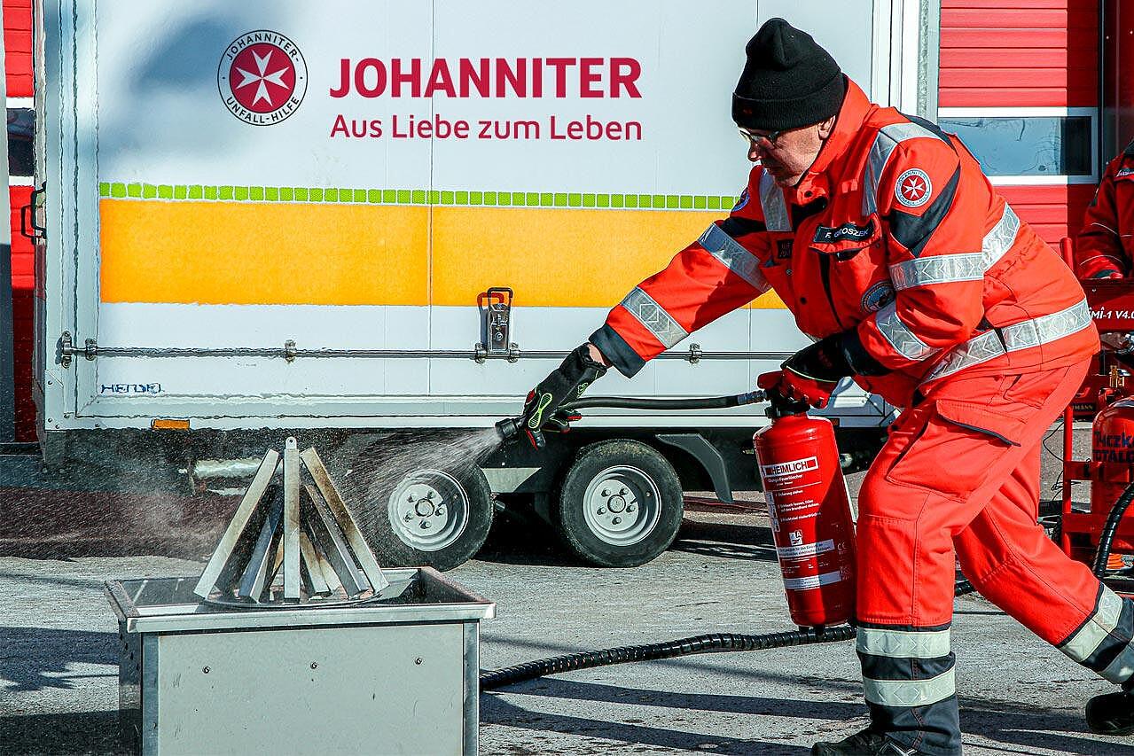 Johanniter in orangefarbener Einsatzkleidung löscht mit Feuerlöscher ein brennendes Übungsgefäß im Brandschutzhelferkurs.
