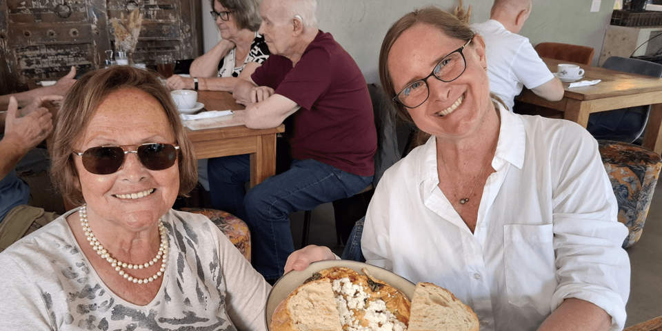 zwei Frauen, die jüngere präsentiert der Kamera eine dekorierten Teller mit Essen. Im Hintergrund weitere Personen an Tischen