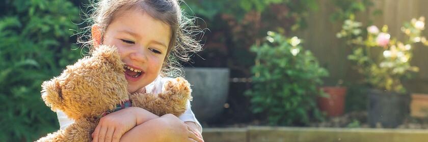 Ein lachendes Kind sitzt im Garten und umarmt fröhlich einen braunen Teddybären im Sonnenschein.