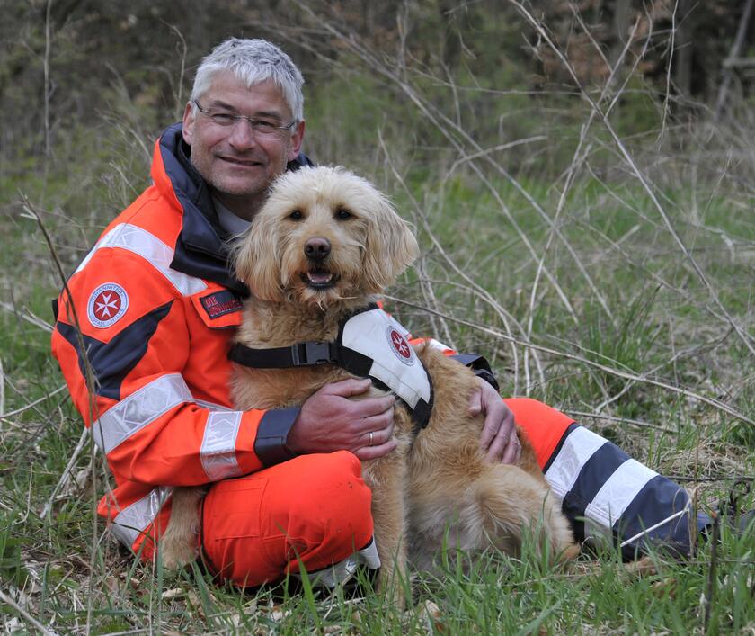 Ein Johanniter-Rettungshundeführer in orangener Einsatzkleidung sitzt in einer Wiese und umarmt seinen freundlichen, lockigen Rettungshund mit weißer Einsatzweste. Beide lächeln in die Kamera und wirken zufrieden und einsatzbereit.
