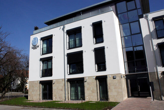 Außenansicht eines drei stöckigem Wohngebäudes mit Flachdach. Die Außenfassade im Erdgeschoss ist mit hellbraunen Steinen verklinkert. der Rest ist weiß