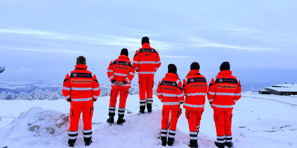 6 Johanniter zu sehen von hinten. Sie schauen von einem schneebedeckten Gipfel ins verschneite Tal.