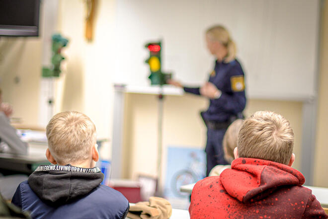 Kinder sitzen an Schultischen und hören einer Frau in Uniform zu, die ihnen gerade etwas erklärt. Neben ihr steht eine kleine Verkehrs-Ampel.