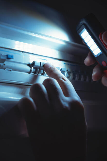 Auf dem Bild ist ein Sicherungskasten zu sehen. Das Licht ist aus, eine Hand mit Taschenlampe leuchtet in den Sicherungskasten und prüft, ob die Sicherungen noch drin sind.