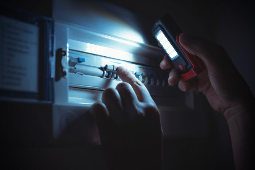 Auf dem Bild ist ein Sicherungskasten zu sehen. Das Licht ist aus, eine Hand mit Taschenlampe leuchtet in den Sicherungskasten und prüft, ob die Sicherungen noch drin sind.